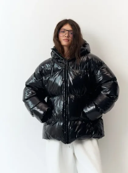 Person posing in a glossy black oversized puffer jacket with white pants, facing the camera wearing glasses.