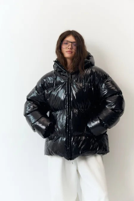 Бомбер жіночий флісовий молочний one size полар-фліс - Person posing in a glossy black oversized puffer jacket with white pants, facing the camera wearing glasses.