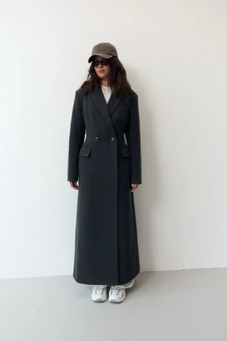 Жіночий графітовий піжак oversize one size - Person in a long dark double-breasted coat, white sneakers, gray cap and sunglasses, against a light wall.