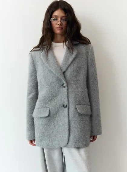 Woman posing in a light gray fuzzy coat and matching pants, wearing rectangular glasses against a white wall.