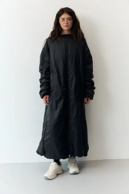 Пуховик жіночий оверсайз екопух до -15°C XS/S M/L - Woman in a long black puffer coat and chunky white sneakers stands against a white wall, facing forward with hands at sides.