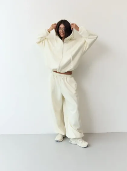 Person wearing an oversized white hoodie and matching sweatpants, standing against a light wall, hands pulling the hood over their head with a smile.