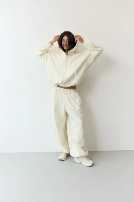 Костюм жіночий oversize молочний з вишивкою футер на флісі - Person wearing an oversized white hoodie and matching sweatpants, standing against a light wall, hands pulling the hood over their head with a smile.