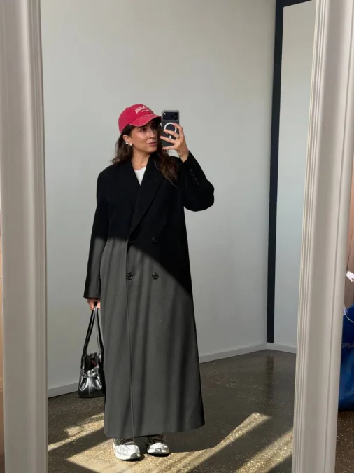Укорочене пальто сіре oversize із вовни - Woman in a long black-to-dark gray color-block coat takes a mirror selfie, wearing a pink cap, white sneakers, and carrying a black handbag.