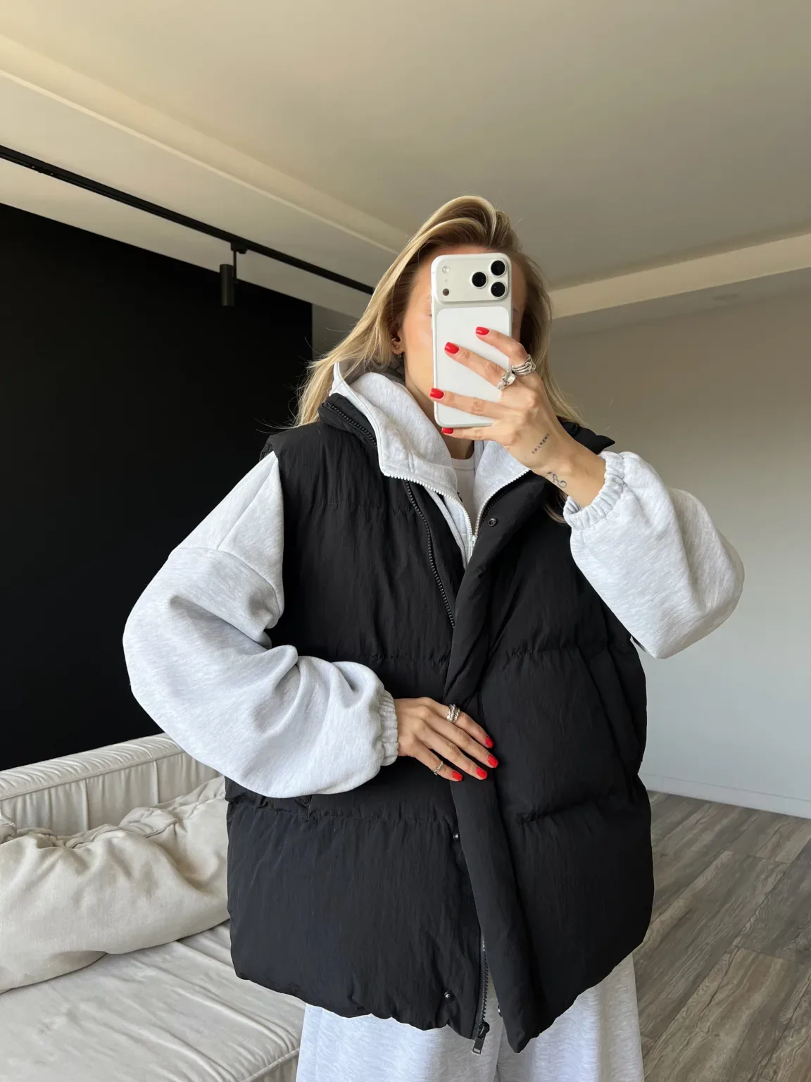 Жіночий верхній одяг: куртки, жилети та бомбери - Person taking a mirror selfie wearing a gray hoodie under a black puffer vest, with red nails and rings, holding a white smartphone.