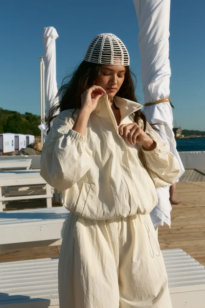 Жіночий верхній одяг: куртки, жилети та бомбери - Woman in a cream outfit and a white woven hat stands on a wooden boardwalk by the sea, adjusting her jacket.