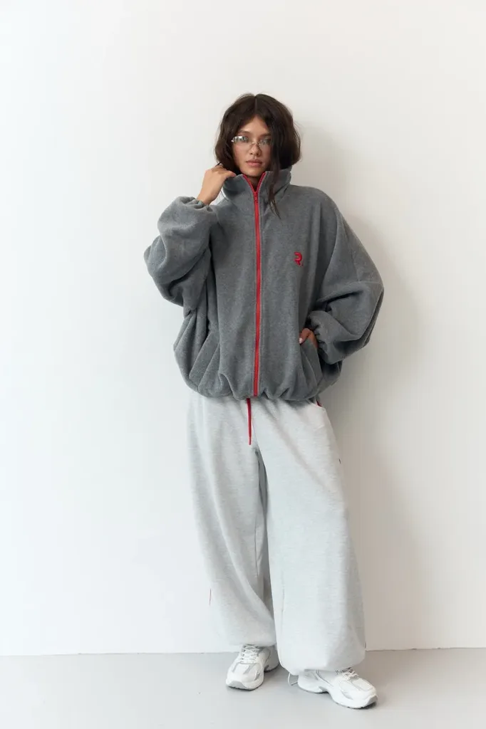 Жіночий верхній одяг: куртки, жилети та бомбери - Person wearing oversized gray zip-front fleece jacket with red trim, paired with wide light gray sweatpants and white sneakers, posing against a white wall.