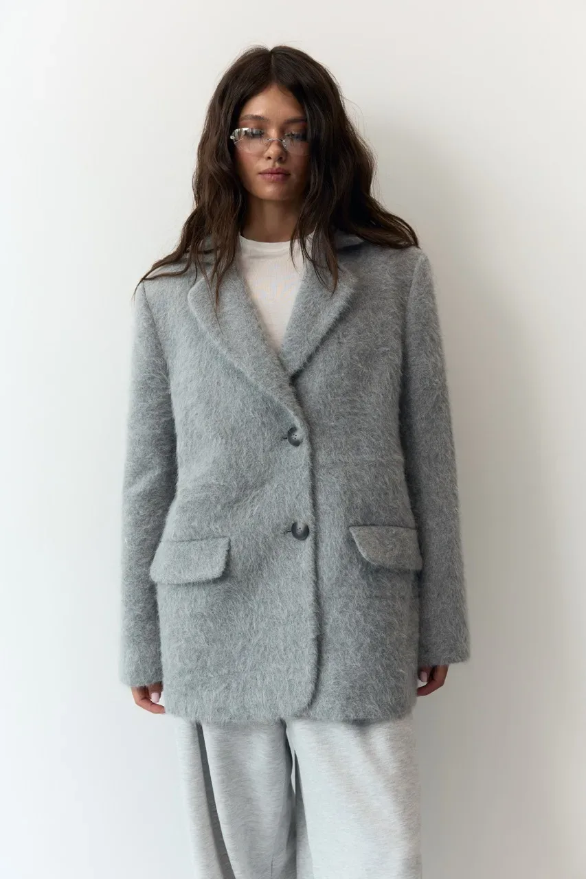 Жіночий верхній одяг: куртки, жилети та бомбери - Woman standing against a white wall in a light gray fuzzy blazer, buttoned once, with a white top and light gray wide-leg pants, wearing rectangular glasses.